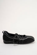 A black low-top lace-up shoe with a matte finish, black laces, and thin white piping details along the side, shown against a plain light gray background.
