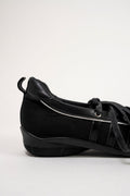 A black low-top sneaker with a mix of suede and smooth leather panels, black laces, and a chunky black sole, shown from a side profile on a light gray background.