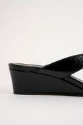 A close-up side view of a black wedge sandal with a glossy strap, set against a plain light gray background.