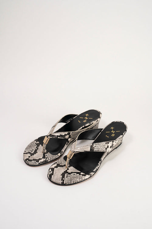 A pair of women's wedge slide sandals featuring a black and beige snakeskin pattern with a single gold-tone thong strap and gold logo details on the insole.