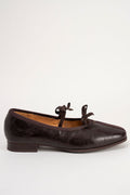 A dark brown leather flat shoe with a pointed toe and two small bow details on the vamp, shown from a side profile against a plain white background.