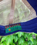 Close-up of a blue and beige mesh trucker hat with a green embroidered patch reading 'LOVEMORE' and 'DON'T BE A PRIDE' with a smiley face, photographed outdoors above green leaves.