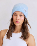 Woman wearing a light blue beanie with a brand logo on a plain background