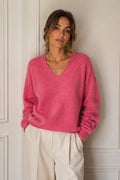 A woman wearing a fuzzy pink V-neck sweater and cream-colored trousers, standing against a white paneled wall.