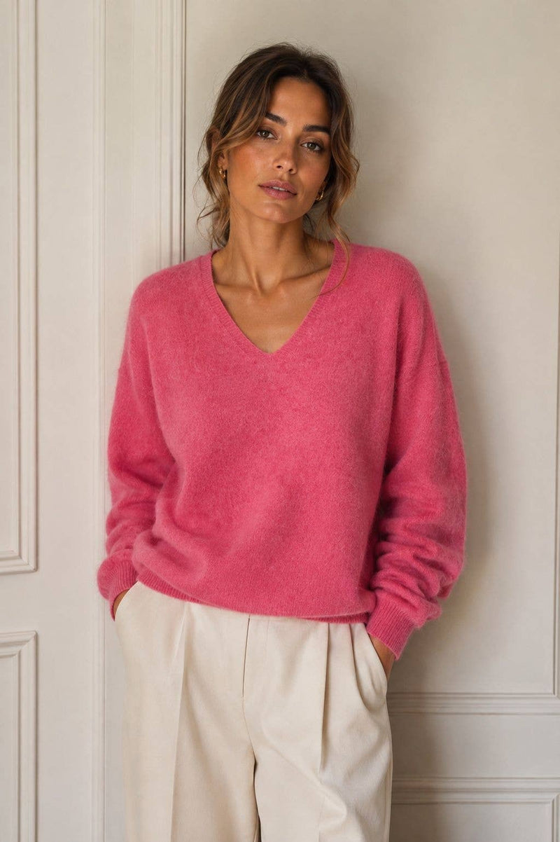 A woman wearing a fuzzy pink V-neck sweater and cream-colored trousers, standing against a white paneled wall.