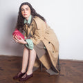 Woman in a beige trench coat holding a pink clutch, sitting on a chair against a white wall.