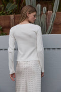 Woman wearing a white ribbed sweater and patterned skirt standing against a neutral background with plants.
