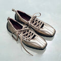 A pair of metallic taupe leather lace-up shoes with a square toe and black soles, displayed on a light gray surface.