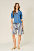 A woman modeling a short-sleeve blue denim shirt with puff sleeves and striped blue, white, and navy shorts, paired with tan woven flats, against a light beige studio background.