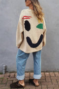 Person wearing a beige sweater with a smiley face design against a gray wall.