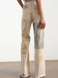 A model from the back wearing high-waisted, wide-leg patchwork trousers in beige, cream, and gray tones with distressed details.