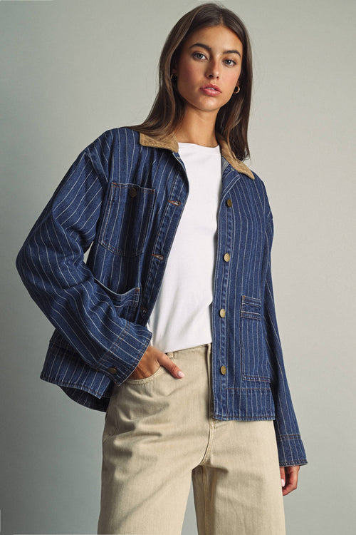 A woman modeling a blue pinstriped denim jacket with a tan corduroy collar, worn over a white t-shirt and beige trousers, against a neutral gray studio background.