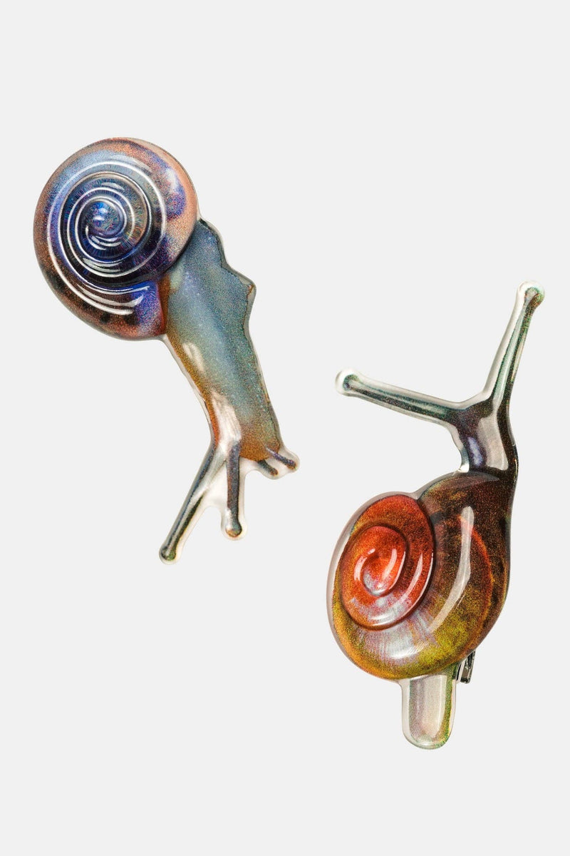 Two decorative snail-shaped hair clips on a plain light gray background. One clip has a blue-purple iridescent shell and a gray body, while the other has a reddish-brown iridescent shell and a gray body.
