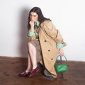 Woman in a beige trench coat holding a green handbag on a wooden floor.