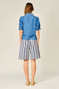 A woman viewed from the back wearing a blue denim short-sleeve jacket and blue-and-white vertically striped shorts, paired with tan pointed flats, against a plain light beige studio background.