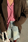 A person wearing an oversized brown blazer over a red and white gingham button-up shirt, paired with light-wash denim shorts and holding a white crochet handbag with red floral details.