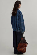 Person wearing a blue denim jacket holding a brown leather bag on a plain background