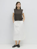 Woman wearing a brown knitted vest over a white shirt with a white skirt on a light gray background