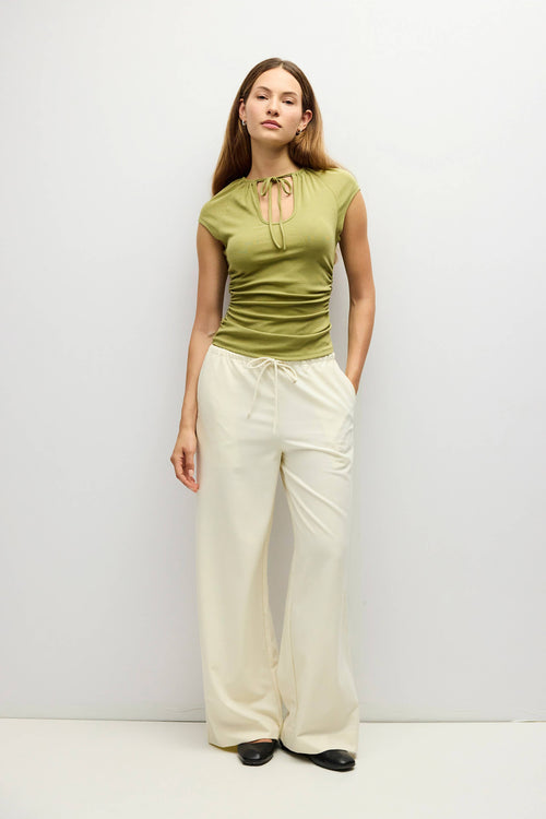 A woman modeling a sleeveless olive green top with a tie-neck detail and cream wide-leg pants, standing against a plain light gray studio background.
