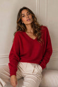 A woman wearing a vibrant red V-neck knit sweater and cream-colored trousers, sitting against a neutral paneled wall.