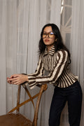 Woman wearing a brown and white striped shirt and black pants, standing indoors with curtains in the background.