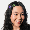 A close-up portrait of a smiling woman with long dark wavy hair wearing two colorful beetle-shaped hair clips, one purple and one green, against a plain light gray background.
